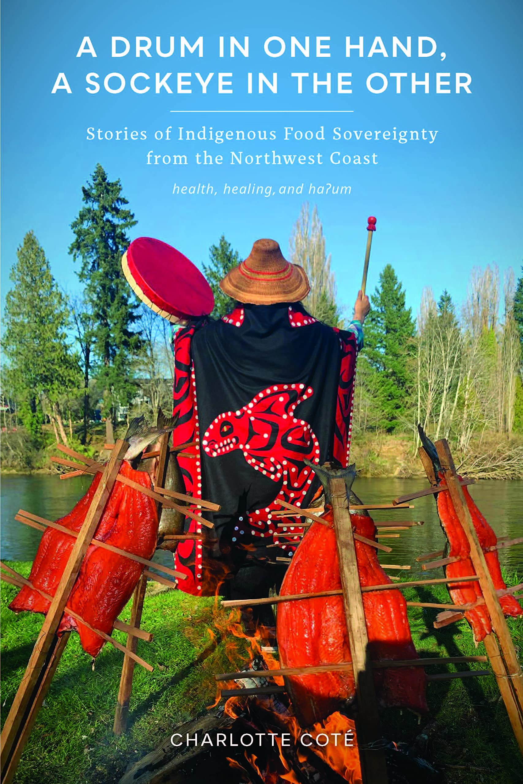 Book cover of A Drum in One Hand, a Sockeye in the Other with traditional salmon racks around a fire and a woman wearing a blanket with salmon embroidered on it and holding her hands in the air, a drum in one hand and a drumstick in the other