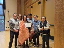 Kai wrapped in a blanket gifted by AIS faculty who stand on both sides of her. She is holding a certificate recognizing her 10 years at UW.