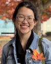 young woman with glasses surrounded by fall foliage with leaves pinned to her jean jacket