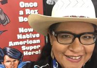 Arigon Starr wearing a cowboy hat, with a poster of her comic in the background.