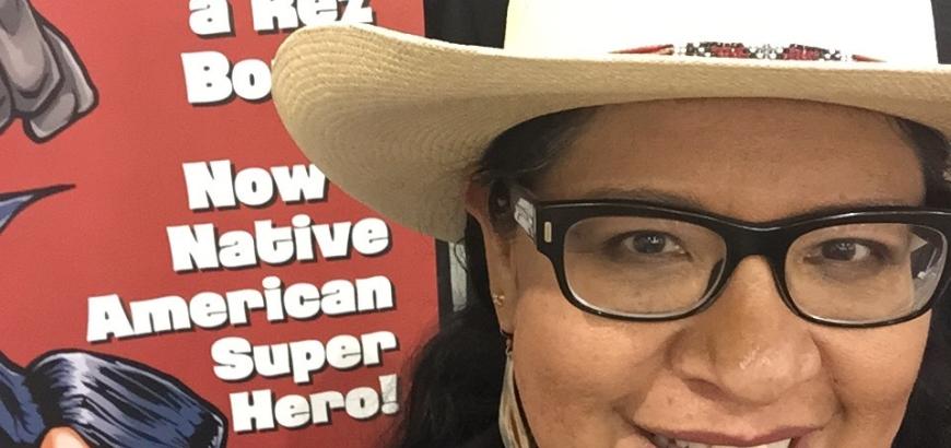 Arigon Starr wearing a cowboy hat, with a poster of her comic in the background.