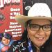 Arigon Starr wearing a cowboy hat, with a poster of her comic in the background.